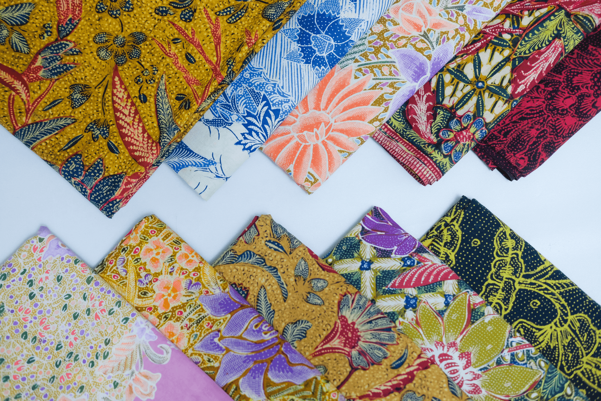 Tumpukan kain batik tulis berbagai warna dan motif bunga, disusun rapi di atas latar putih